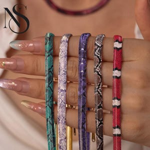 May include: Five flat herringbone chain necklaces featuring various snake skin patterns in teal, light purple, dark purple, brown, and a red, white, and black striped design. Each metal chain has a smooth, reflective finish.