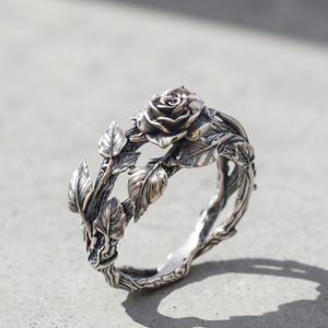 May include: A silver ring featuring a detailed three-dimensional rose bloom as the centerpiece, with sculpted leaves and thorny vine textures wrapping around the entire band, finished with a polished metallic sheen and dark oxidized accents.