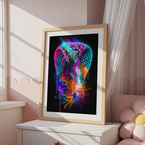 Könnte beinhalten: Ein gerahmter Wandkunstdruck mit einer lebendigen, abstrakten Illustration einer Kobra. Die Schlange ist mit neonblauen, pinken, orangen und gelben Farbspritzern vor einem schwarzen Hintergrund dargestellt und befindet sich in einem hellen Holzrahmen.