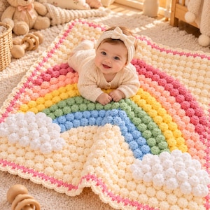 May include: A square crochet baby blanket featuring a textured bobble stitch rainbow design in pink, yellow, green, and blue. The blanket includes white bobble stitch clouds at the ends of the rainbow and a cream-colored background with a pink border.