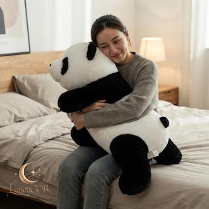 May include: A large, plush panda stuffed animal featuring a soft white body with black patches around the eyes, black ears, and black limbs. The toy has a rounded head, a small black nose, and a short tail, crafted from fuzzy material.