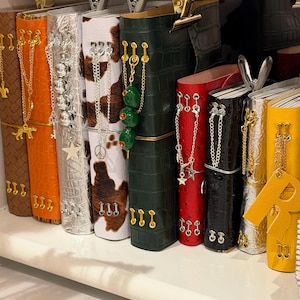 May include: A row of vertical journals with textured covers in brown, orange, silver, cow print, dark green, red, black, and yellow. Each journal features metal eyelets, gold or silver chains, and charms including stars, a peace sign, and olives.