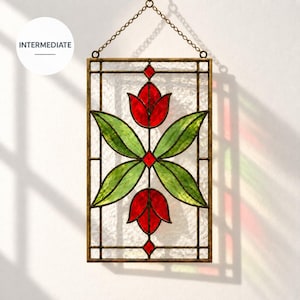 Op de afbeelding: Een rechthoekige glas-in-lood zonnevanger met twee rode tulpbloemen en vier groene bladeren tegen een achtergrond van getextureerd helder glas. Het stuk is gevat in een donkere metalen rand en hangt aan een bijpassende metalen ketting.