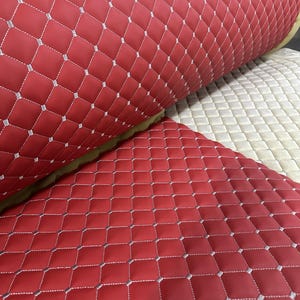 May include: A roll of red synthetic leather fabric featuring a quilted diamond pattern with silver stitching. The material has a smooth, padded texture and is displayed alongside a section of beige quilted fabric with matching silver stitching.