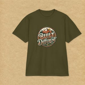 Op de afbeelding: Een olijfgroen t-shirt met korte mouwen en een ronde afbeelding op de borst. Het ontwerp toont een bergketen met dennenbomen en een zon, met de tekst "BUILT DIFFERENT" in een gestileerd, gelaagd lettertype in het midden van het embleem.