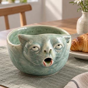 Peut inclure: Un bol en céramique avec un vernis vert clair brillant, sculpté en forme de visage de chat avec des yeux grands et expressifs et une bouche ouverte et circulaire. Le bol présente une surface texturée et un bord arrondi pour contenir des objets.