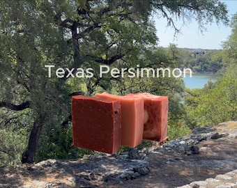 May include: Three rectangular bars of soap arranged in a row, featuring varying shades of reddish-brown, light peach, and deep terracotta. The text "Texas Persimmon" is displayed in white lettering above the soap bars against a natural outdoor backdrop.