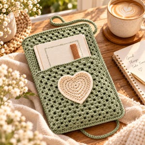 May include: A sage green crocheted pouch featuring a granny square pattern and a cream-colored crocheted heart appliqué on the front pocket. The pouch includes a matching green cord handle and contains a small notebook with a brown spine.