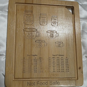 May include: A rectangular bamboo cutting board featuring laser-engraved kitchen conversion charts for dry weights and liquid volumes, illustrations of jars and measuring cups, and the text Not Food Safe at the bottom edge.