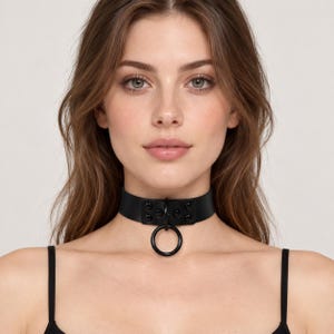 Könnte beinhalten: Ein schwarzes Halsband mit einem glatten, breiten Band und einem zentralen Ringelement. Das Design umfasst einen verstärkten Vorderteil, der mit vier kleinen schwarzen Nieten befestigt ist und für ein einheitliches Erscheinungsbild sorgt.