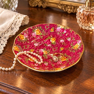 May include: A round ceramic trinket dish featuring a vibrant magenta background with a detailed pattern of yellow and pink roses. The dish has a scalloped edge with a gold-toned rim. A string of white pearls and a gold ring rest inside the dish.