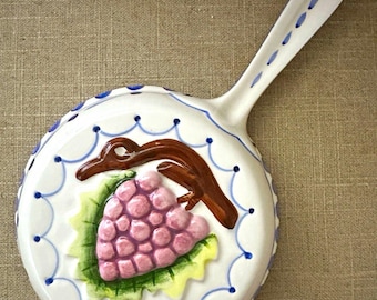 Può includere: Una padella decorativa in ceramica bianca con un rilievo tridimensionale raffigurante un grappolo d'uva rosa, foglie verdi e un tralcio marrone. Il bordo e il manico sono decorati con un motivo a festoni blu e puntini blu.