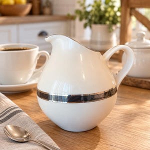 May include: A cream-colored ceramic creamer pitcher with a rounded body, a curved handle, and a pouring spout. The pitcher features a horizontal band of dark gray or black with thin silver metallic trim encircling the middle section.