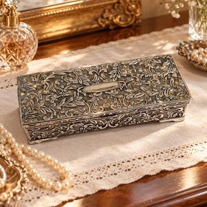May include: A rectangular silver-toned metal box featuring an intricate embossed floral and leaf pattern across the lid and sides. The lid includes a smooth, blank oval plaque in the center for personalization, and the box rests on four small feet.