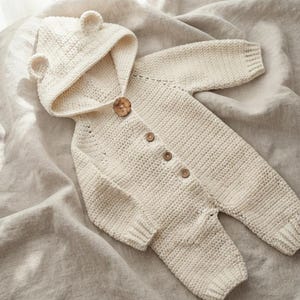 May include: A cream-colored, long-sleeved, hooded crochet romper featuring two small bear-like ears on the hood. The garment is fastened down the front with four round, brown wooden buttons and has ribbed cuffs at the wrists and ankles.