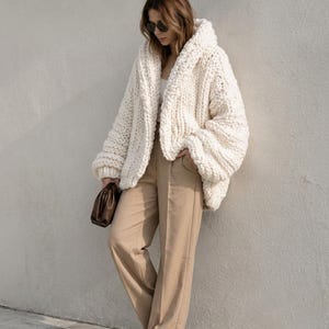 Può includere: Un cardigan oversize in maglia grossa color bianco sporco con colletto ampio e maniche lunghe, abbinato a pantaloni beige a vita alta, un top bianco, scarpe da ginnastica bianche e una piccola pochette marrone in mano.