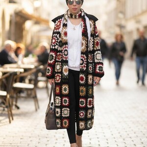 May include: A long, open-front crochet cardigan featuring a multi-coloured granny square pattern in black, white, red, and olive green. The garment is worn over a plain white t-shirt and black leggings, paired with a matching crochet scarf and a handbag.
