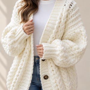 Puede incluir: Un cárdigan de punto grueso en color blanco roto con un patrón de trenzas prominente, mangas largas abullonadas con puños acanalados y un frente abotonado con dos botones visibles de color marrón oscuro sobre una camiseta blanca y jeans azules.