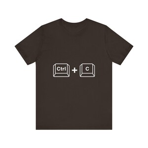 Pode incluir: Uma t-shirt de manga curta castanho-escura com uma impressão gráfica branca no peito. O design apresenta dois ícones quadrados de teclas de teclado com o texto Ctrl e C no interior, separados por um sinal de mais, para o comando copiar.