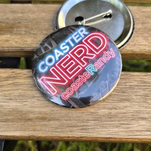 Könnte beinhalten: Ein runder Ansteckbutton mit schwarzem Hintergrund und einer dunklen Grafik einer Achterbahnstruktur. Der Text lautet COASTER NERD in fetten weißen und roten Buchstaben, mit dem kleineren Text coasterRandy in rosa Schreibschrift darunter.