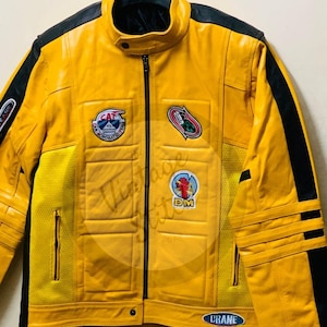 May include: A bright yellow jacket with black shoulder and side panels, featuring a stand-up collar, a central zipper, and multiple embroidered patches on the chest. The patches include text reading CAT, DM, and CRANE, with quilted detailing.