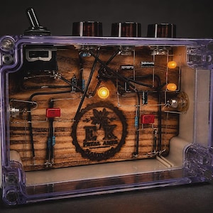 May include: A transparent effects pedal enclosure featuring a wooden interior panel with the laser-engraved text EK PETAL AMPS HANDMADE. The device includes three dark knobs, a black toggle switch, internal wiring, and two glowing amber lights.