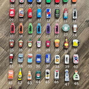 May include: A collection of fifty small, colorful beverage-themed shoe charms arranged in a grid. The charms feature various soda cans, energy drinks, coffee cups, beer bottles, and alcoholic beverage containers, each labeled with a number from 01 to 50.