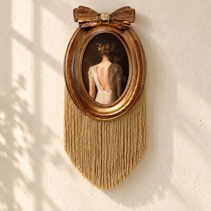 May include: An oval-shaped wall frame with a metallic bronze finish, topped with a decorative bow. The frame contains a portrait of a person seen from behind in a white dress. Long, golden-colored fringe hangs from the bottom of the frame.
