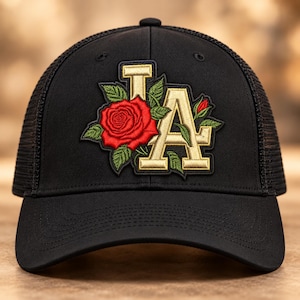 May include: A black trucker hat featuring an embroidered patch on the front. The patch displays the letters LA in gold with a large red rose and green leaves intertwined around the letters. The hat has a solid front panel and mesh back.