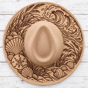 May include: A tan wide-brimmed hat featuring a dark brown line art illustration on the brim. The design depicts ocean waves, tall beach grass, a large scallop shell, a sand dollar, and various spiral seashells arranged around the crown.