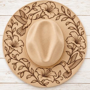 May include: A tan felt fedora hat featuring a dark brown laser-engraved design on the brim. The pattern consists of two hummingbirds and large hibiscus flowers connected by leafy vines that encircle the crown of the hat.