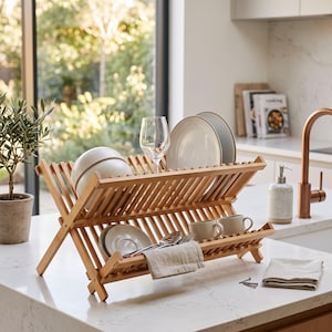 Può includere: Uno scolapiatti pieghevole in bambù a due livelli con design a doghe. Il supporto contiene diversi piatti in ceramica, ciotole, due tazze, un bicchiere trasparente e posate in metallo, con un panno beige piegato sul bordo anteriore.
