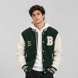 Op de afbeelding: Een donkergroen varsity-jack met crèmekleurige mouwen, een witte letter B-patch op de borst en de tekst Better Days Ahead in het wit geborduurd. Het jack wordt gedragen over een witte hoodie met witte trekkoorden en drukknopen.