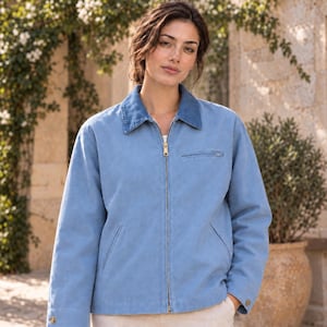 May include: A light blue zip-up jacket featuring a contrasting darker blue collar, a gold-toned metal front zip, a single horizontal zipped chest pocket, two side pockets, and buttoned cuffs on the long sleeves, constructed from a solid fabric.