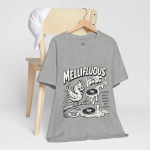 May include: A heather grey short-sleeve t-shirt featuring a black and white graphic of a bird on a branch with vinyl records and the text "MELLIFLUOUS" and "A sound that is sweet, smooth and musical" along with "BEAUTIFUL WORDS CLUB".