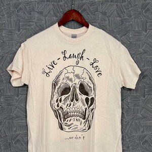 May include: A white t-shirt with a black and white illustration of a skull with the text "Live - Laugh - Love... or don't"