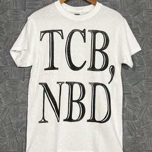 Może przedstawiać: Biała koszulka z czarnym nadrukiem "TCB, NBD".