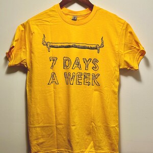 May include: A yellow t-shirt with a black graphic of a rolled-up cigarette with flames on each end. The text "7 DAYS A WEEK" is printed below the graphic.