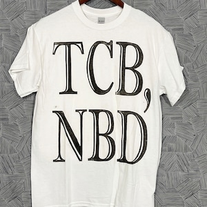 Może przedstawiać: Biała koszulka z czarnym nadrukiem "TCB, NBD".