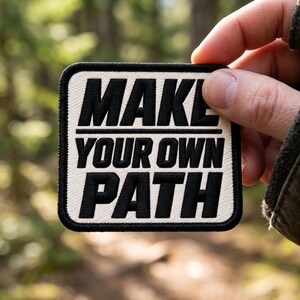 May include: A square embroidered patch with a cream-colored background and a thick black border. The patch features bold black text that reads "MAKE YOUR OWN PATH" in three stacked lines, with a horizontal line separating the first two words.