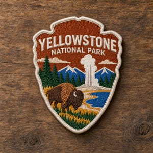 May include: A shield-shaped embroidered patch featuring the text YELLOWSTONE NATIONAL PARK in white. The design depicts a brown bison in a grassy field, a blue geyser plume, blue mountains with white peaks, and green pine trees on a rust background.