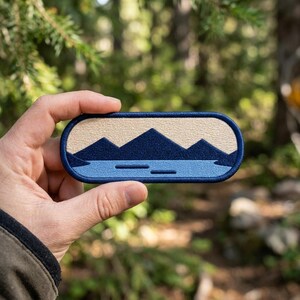 May include: An oval-shaped embroidered patch featuring a landscape design. The patch has a dark blue border, a cream-colored sky, dark blue mountain silhouettes, and a light blue water section with two small horizontal lines.