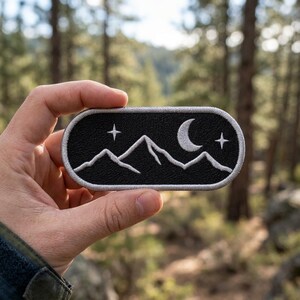 May include: An oval-shaped embroidered patch featuring a black background with white stitched mountain peaks, a crescent moon, and two four-pointed stars. The patch is finished with a light gray stitched border around the entire edge.