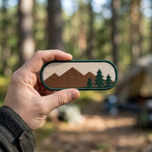 May include: An oval-shaped embroidered patch featuring a landscape design. The patch displays brown mountain peaks against a cream-colored sky, with three dark green pine trees positioned on the right side, all enclosed by a dark green border.