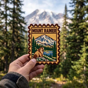 May include: A rectangular embroidered patch with a scalloped border featuring a graphic of Mount Rainier, a marmot, a lake, and evergreen trees. The text MOUNT RAINIER is printed in dark brown at the top of the yellow and brown design.