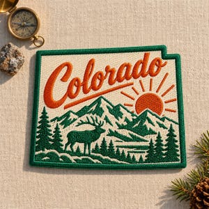 May include: A rectangular embroidered patch featuring the word Colorado in orange script, a rising orange sun, green mountain peaks, and a green elk silhouette against a cream background, bordered by a thick dark green frame.