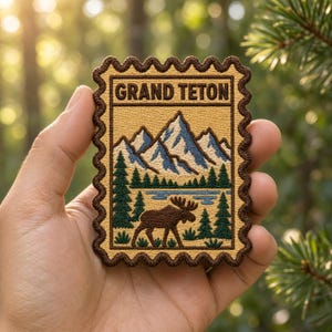 May include: A rectangular embroidered patch shaped like a postage stamp with a dark brown scalloped border. The design features the text GRAND TETON above a landscape of snow-capped blue mountains, a blue lake, green pine trees, and a brown moose.