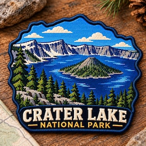 May include: A blue embroidered patch featuring a landscape of Crater Lake National Park with snow-capped mountains, a deep blue lake, a central island, and evergreen trees. The text CRATER LAKE NATIONAL PARK is stitched in white and yellow.