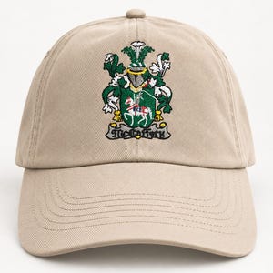 May include: A beige baseball cap featuring an embroidered coat of arms on the front panel. The design includes a green shield with a white horse, a knight's helmet, and the text McCaffrey on a scroll banner at the base of the emblem.