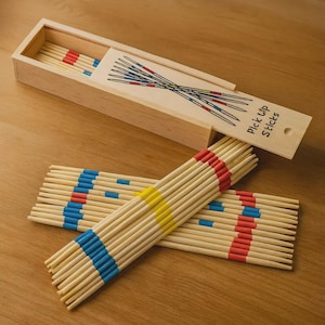 Op de afbeelding: Een houten doos met een schuifdeksel met de tekst Pick Up Sticks en een afbeelding van gekruiste stokjes. In en buiten de doos liggen talloze lichtgekleurde houten stokjes, elk gemarkeerd met banden van rode, blauwe en gele verf op verschillende afstanden.